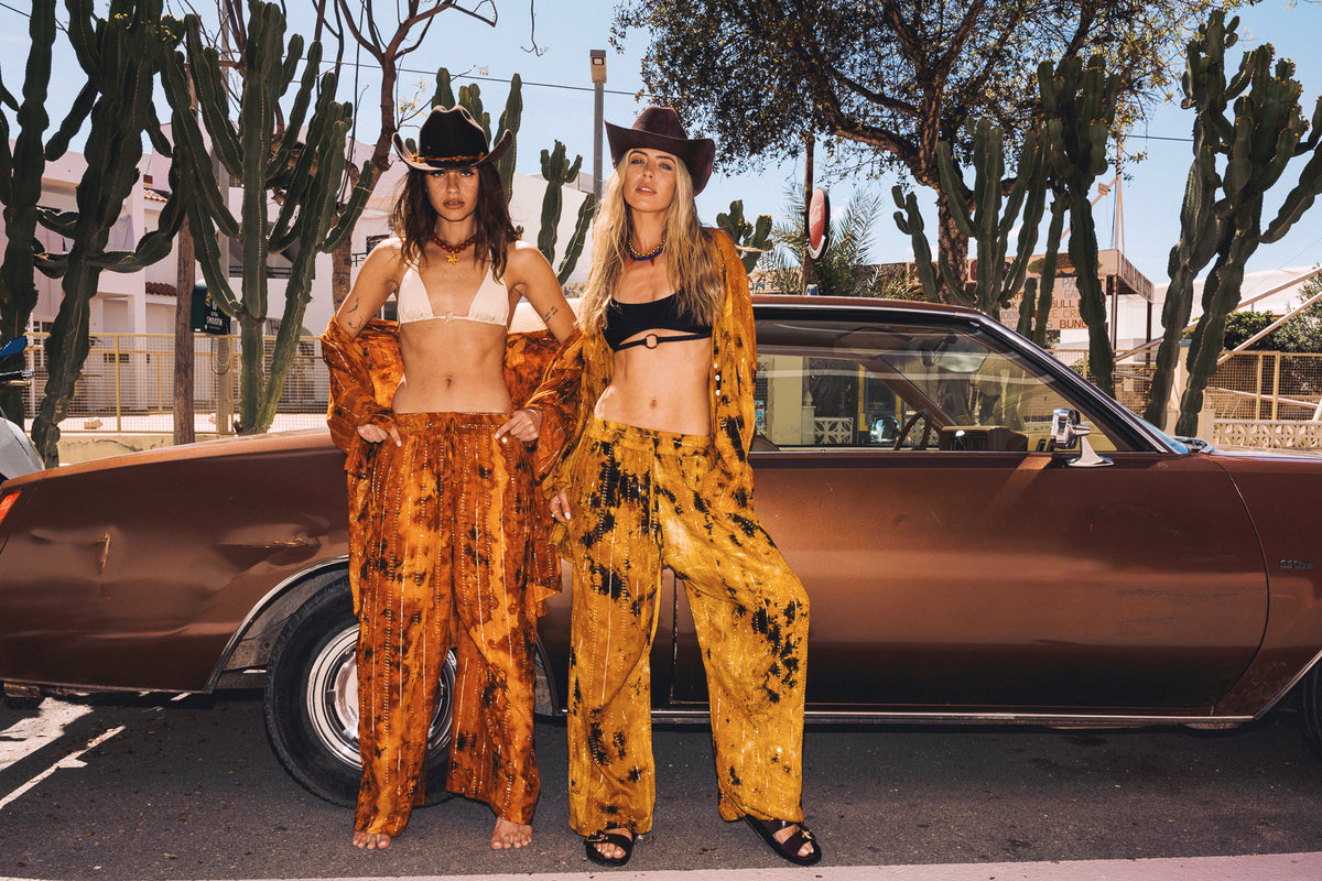 Two women in matching outfits standing next to a vintage car with cacti in the background.